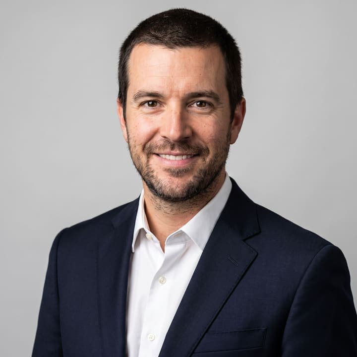 The same person in a smart-casual startup headshot with an office blur